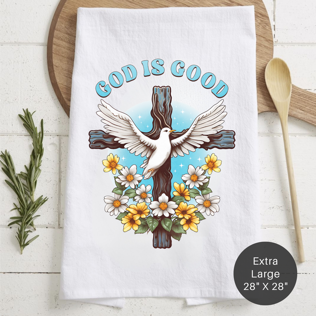 White tea towel with 'God is Good' text, dove on cross, and flowers on a wooden surface.
