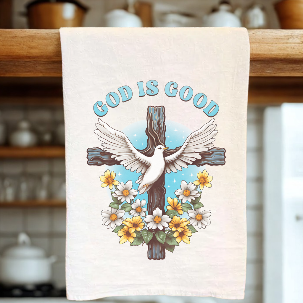 Tea towel with 'God is Good' text, dove, cross, and flowers on a wooden rack.