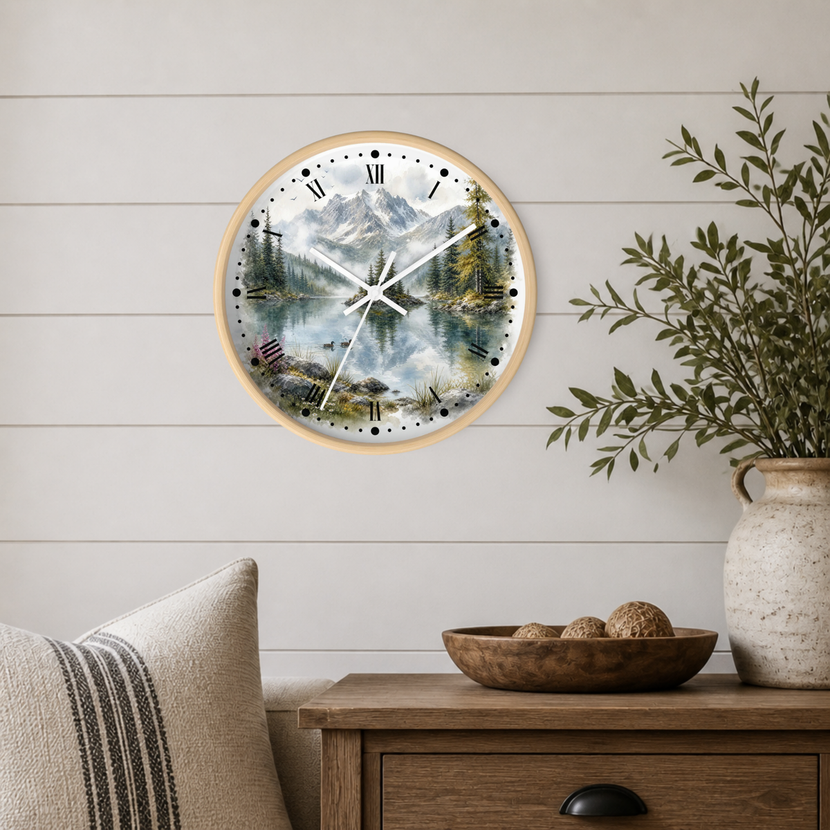 A round wall clock with a light wood frame and a scenic mountain lake print, mounted on a white shiplap wall above a wooden side table with a vase and bowl.