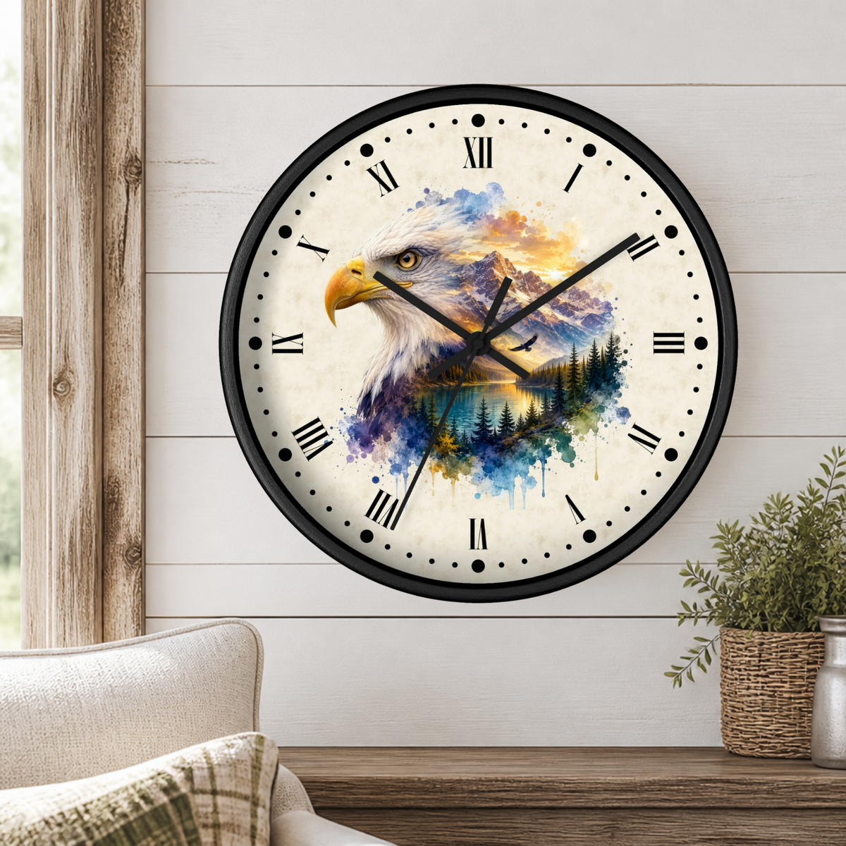 A round wall clock with a black frame and Roman numerals, featuring a watercolor-style bald eagle over a mountain and lake landscape on the face. The clock is mounted on a white shiplap wall above a wooden table with a potted plant and part of a sofa visible.