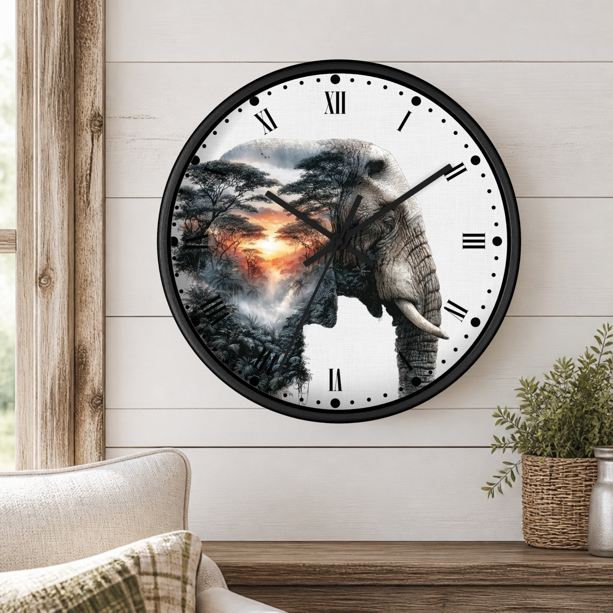A round wall clock with a black frame and Roman numerals, featuring a printed design of an elephant silhouette blending into a savanna landscape with a sunset. The clock is mounted on a white wood-paneled wall above a wooden table with a potted plant and a sofa visible.