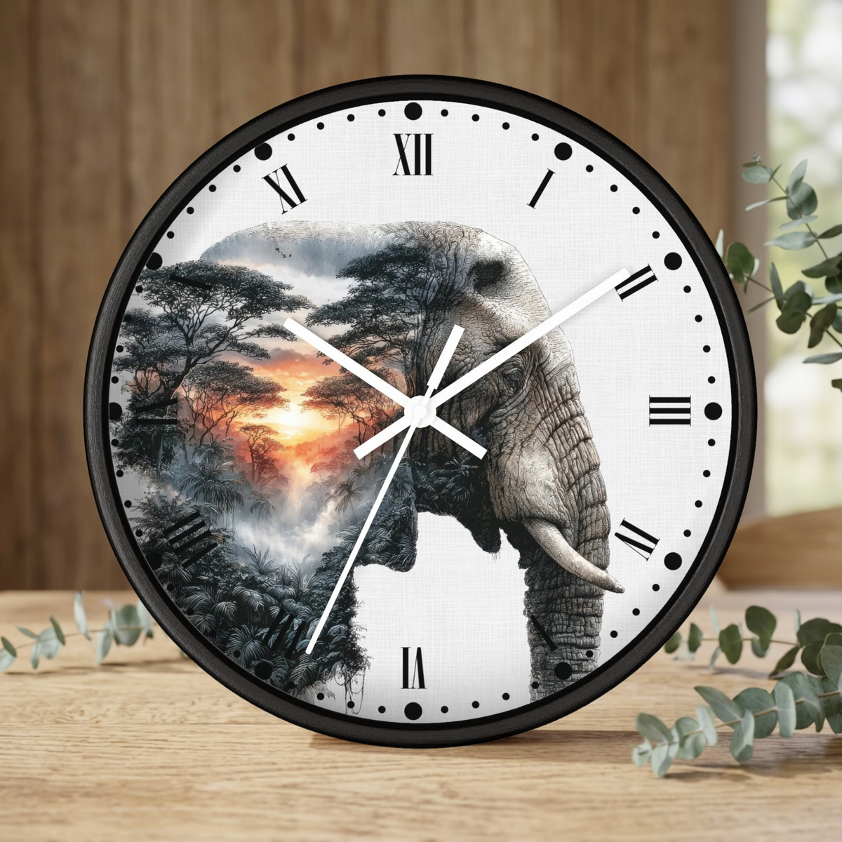 Round wall clock with a black frame and a white face featuring a large elephant and tree landscape design, styled on a wooden surface with eucalyptus leaves.