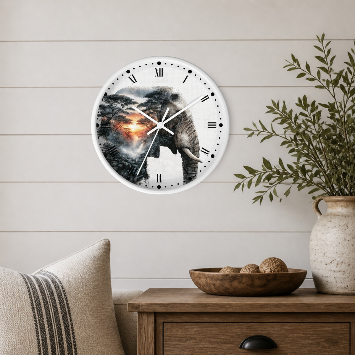 A round wall clock with a white frame and a printed elephant and forest scene, mounted on a light wood paneled wall above a wooden side table with a vase and bowl.