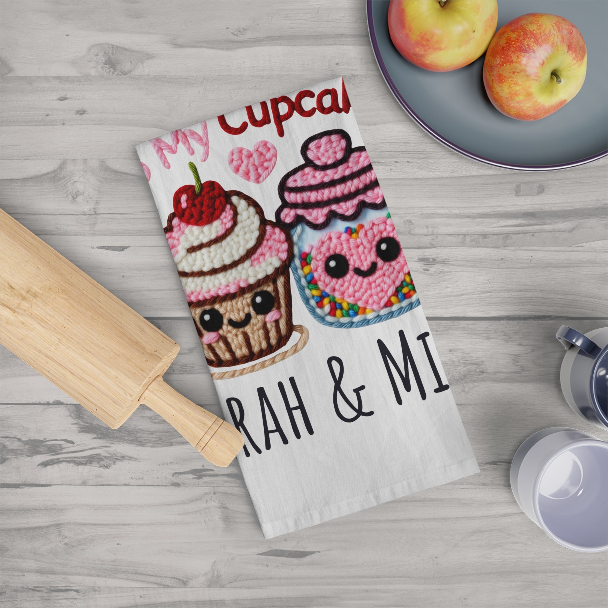 Kawaii-style cupcake-themed kitchen towel on a wooden surface with apples and a rolling pin.