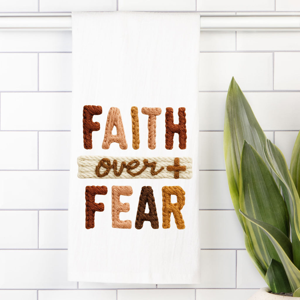 White tea towel text 'FAITH over FEAR' made of yarn on a white tiled wall with a plant.