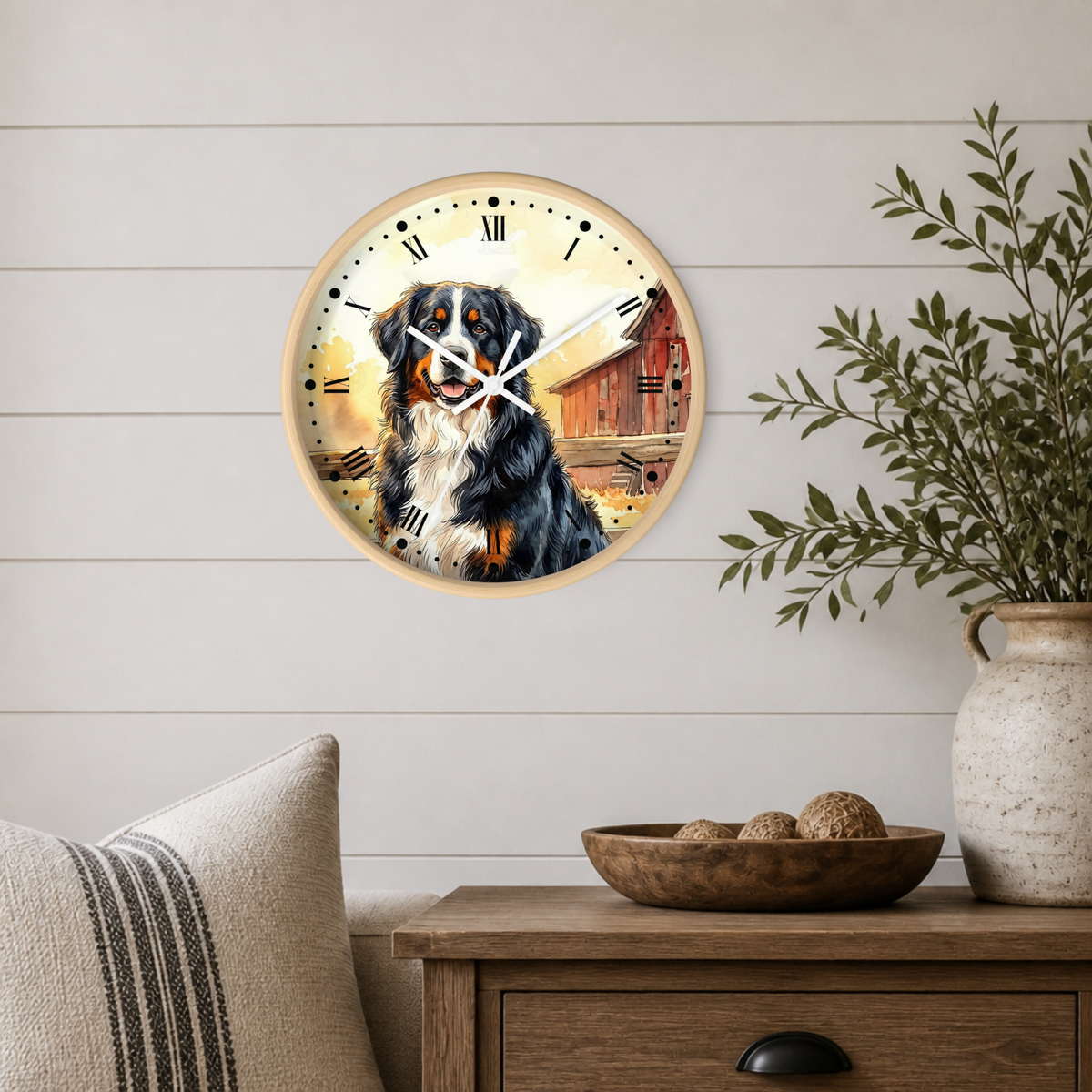 Round wall clock with a light wood frame featuring a painted Bernese mountain dog and barn scene, displayed on a white shiplap wall above a wooden side table with a plant and decorative bowl.