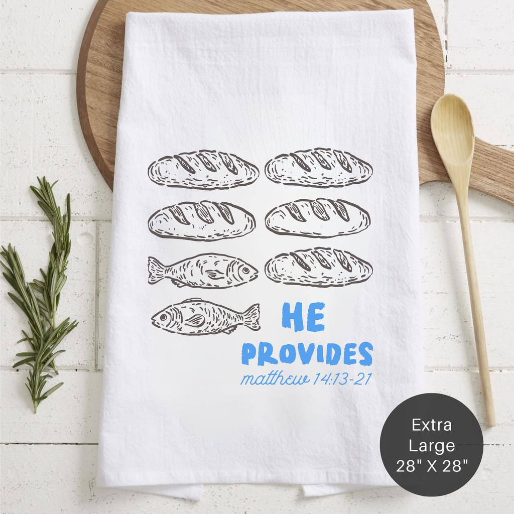 White towel with bread and fish illustrations and 'HE PROVIDES' text on a wooden surface.