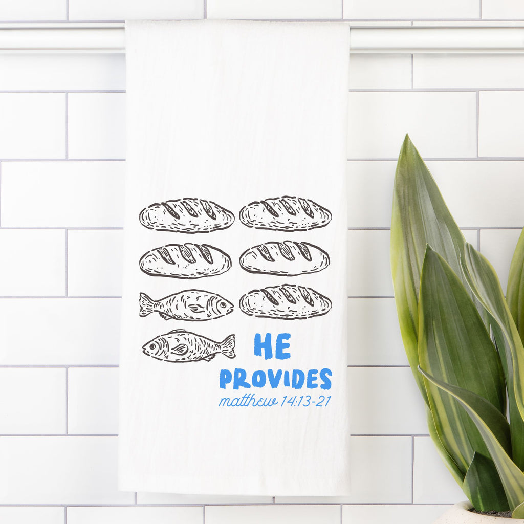 Tea towel with bread and fish illustrations and 'He Provides' text on a tiled wall.