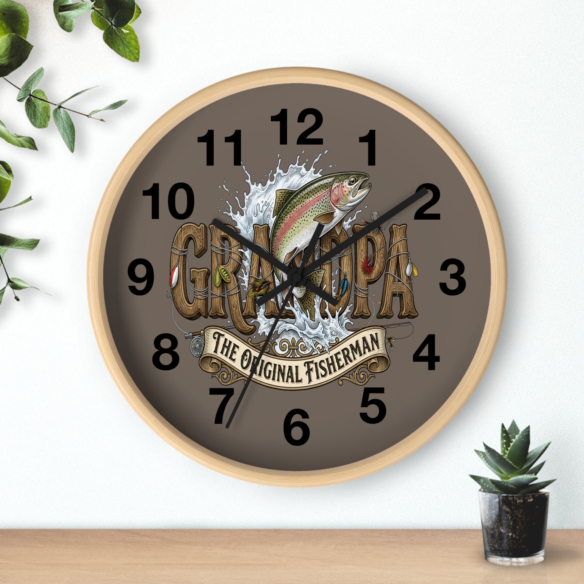 Wall clock with a fish design and 'Grillocra' text on a brown background, placed on a wooden surface with plants.