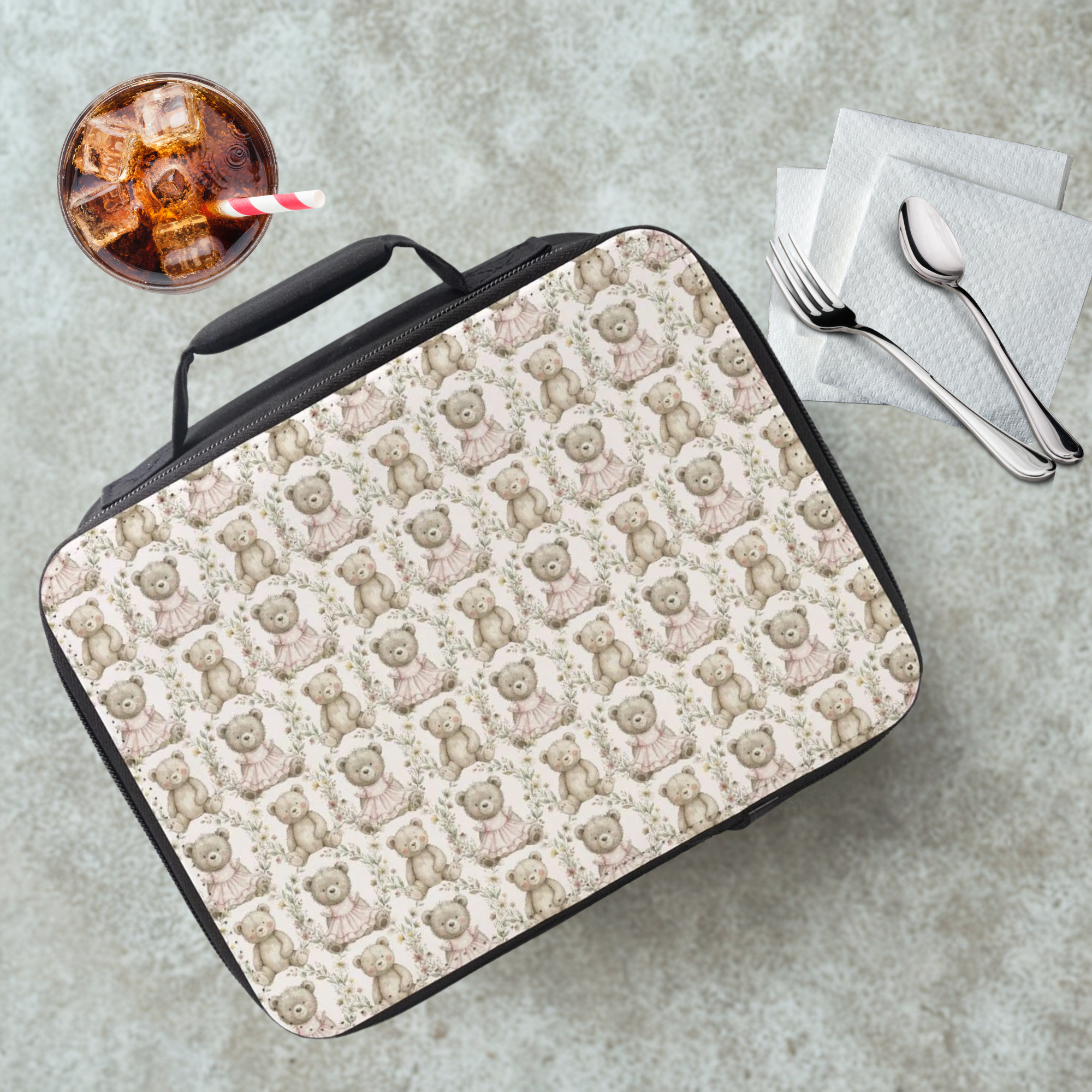 Lunch bag with teddy bear pattern on a concrete surface with a drink and utensils.