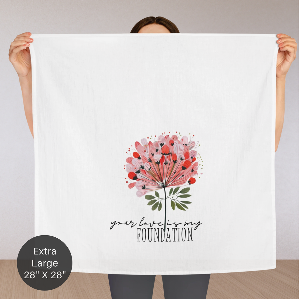 Person holding an extra large white tea towel with a floral design and text 'your love is my foundation' on a plain background