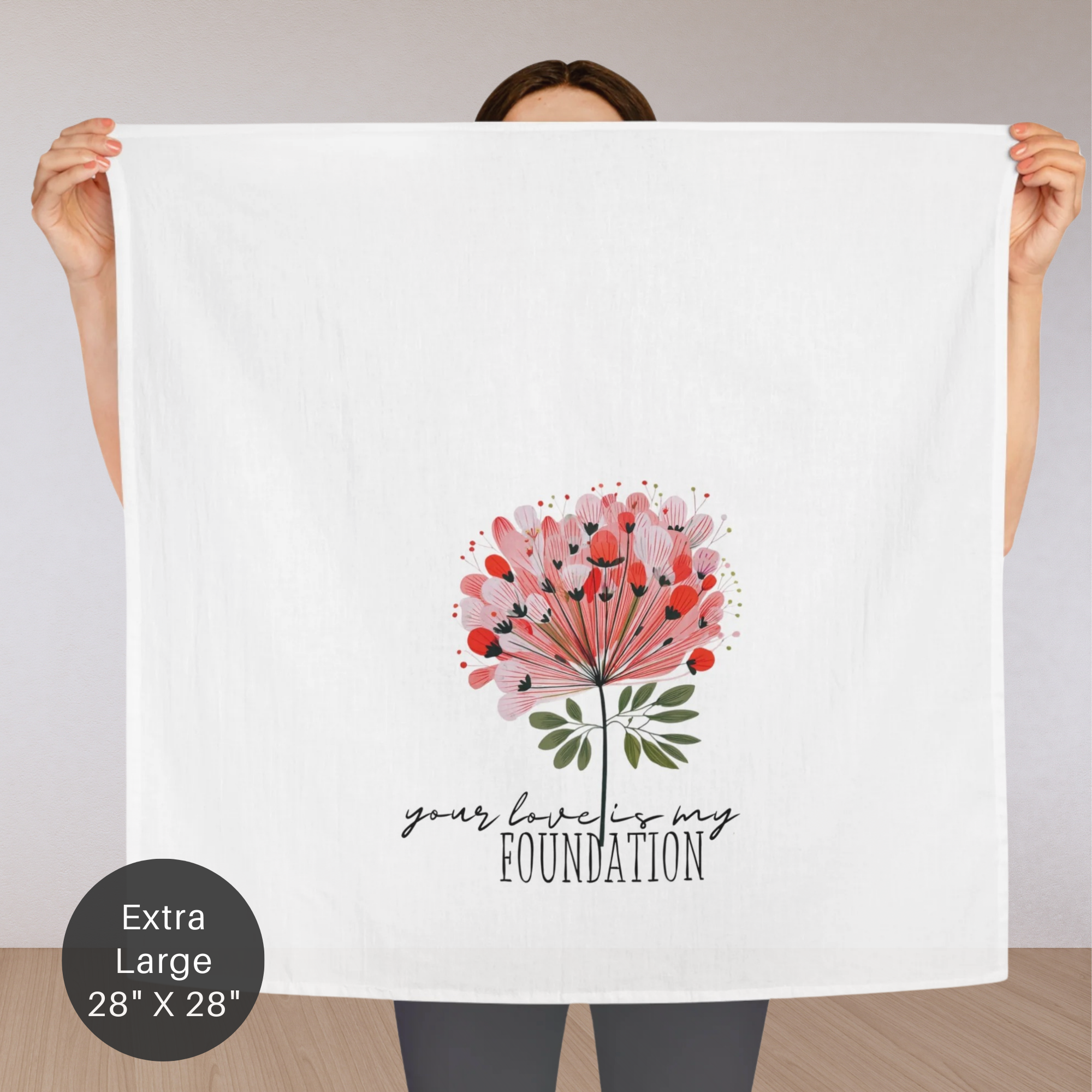 Person holding an extra large white tea towel with a floral design and text 'your love is my foundation' on a plain background