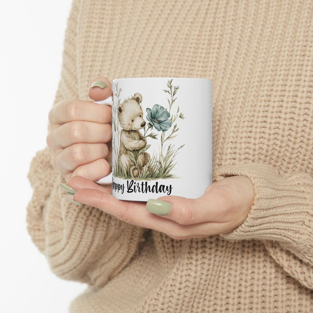 Person holding a mug with a bear and floral design, text 'Happy Birthday', wearing a beige sweater.