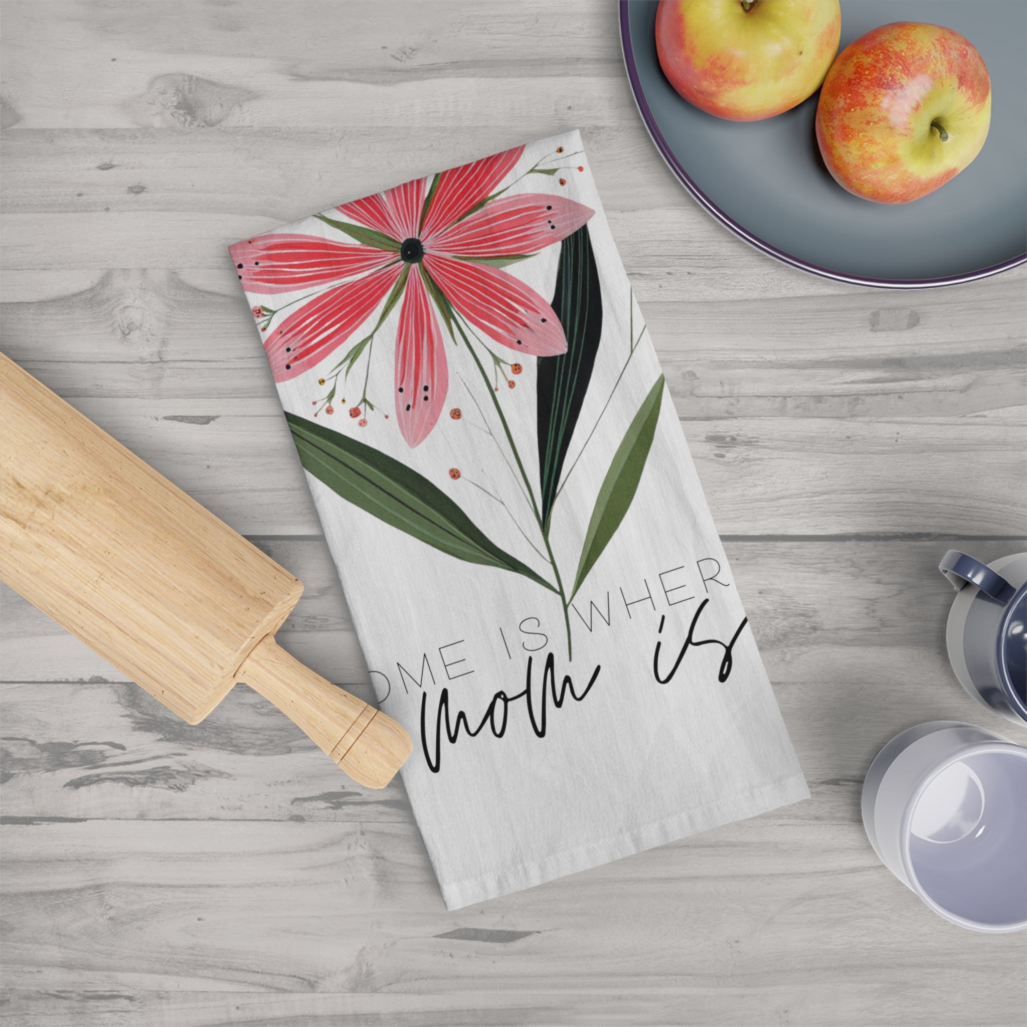 Floral tea towel with text 'Home Is Where Mom Is' on a kitchen counter with apples and a rolling pin.
