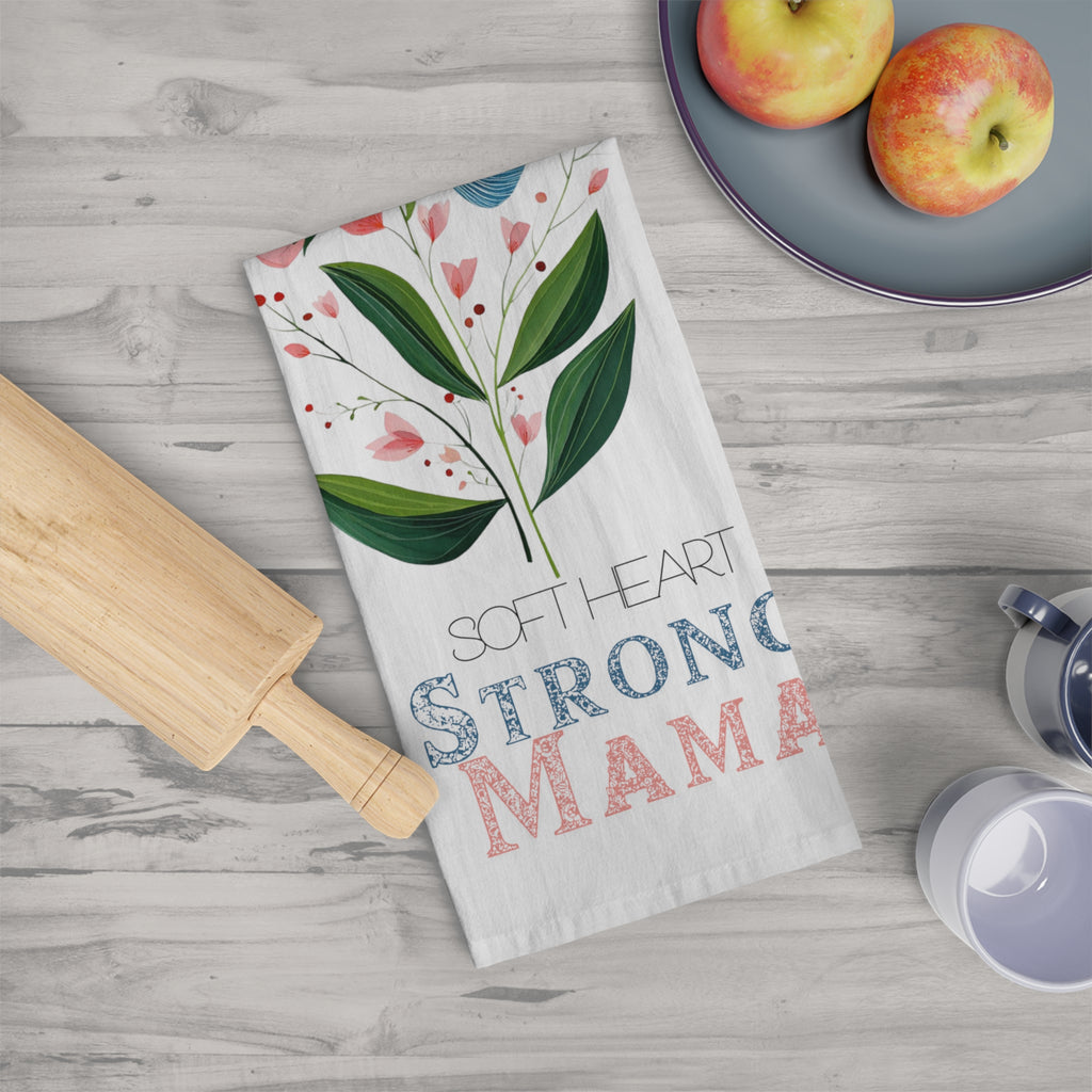 Folded kitchen towel with floral design and text on a wooden surface with apples and a rolling pin.
