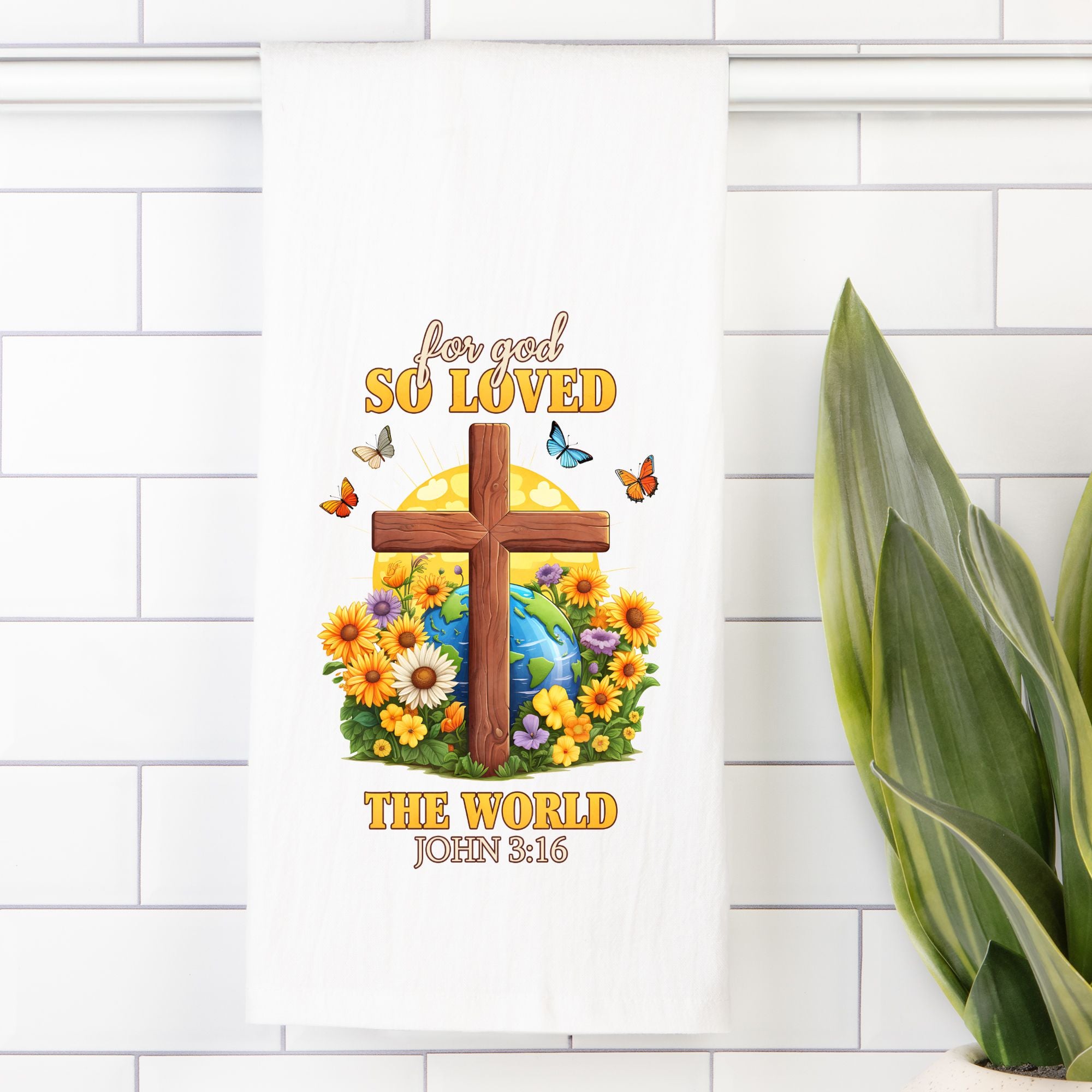 Decorative tea towel with cross, flowers, and 'For God So Loved the World' text on a white tiled wall.