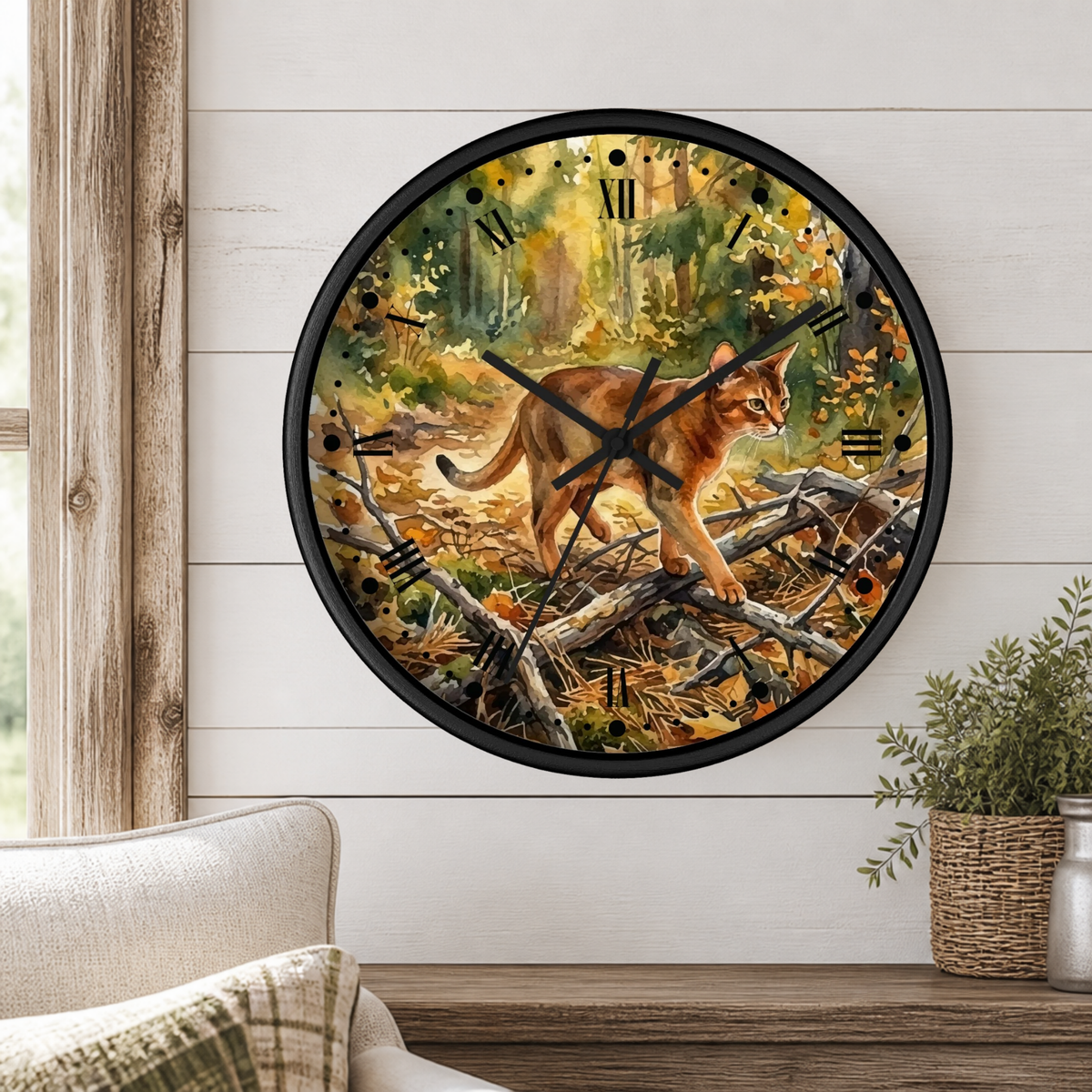 A round wall clock with a black frame and a printed watercolor  forest scene featuring a walking cat, mounted on a white shiplap wall above a wooden mantel with a plant and part of a chair visible.