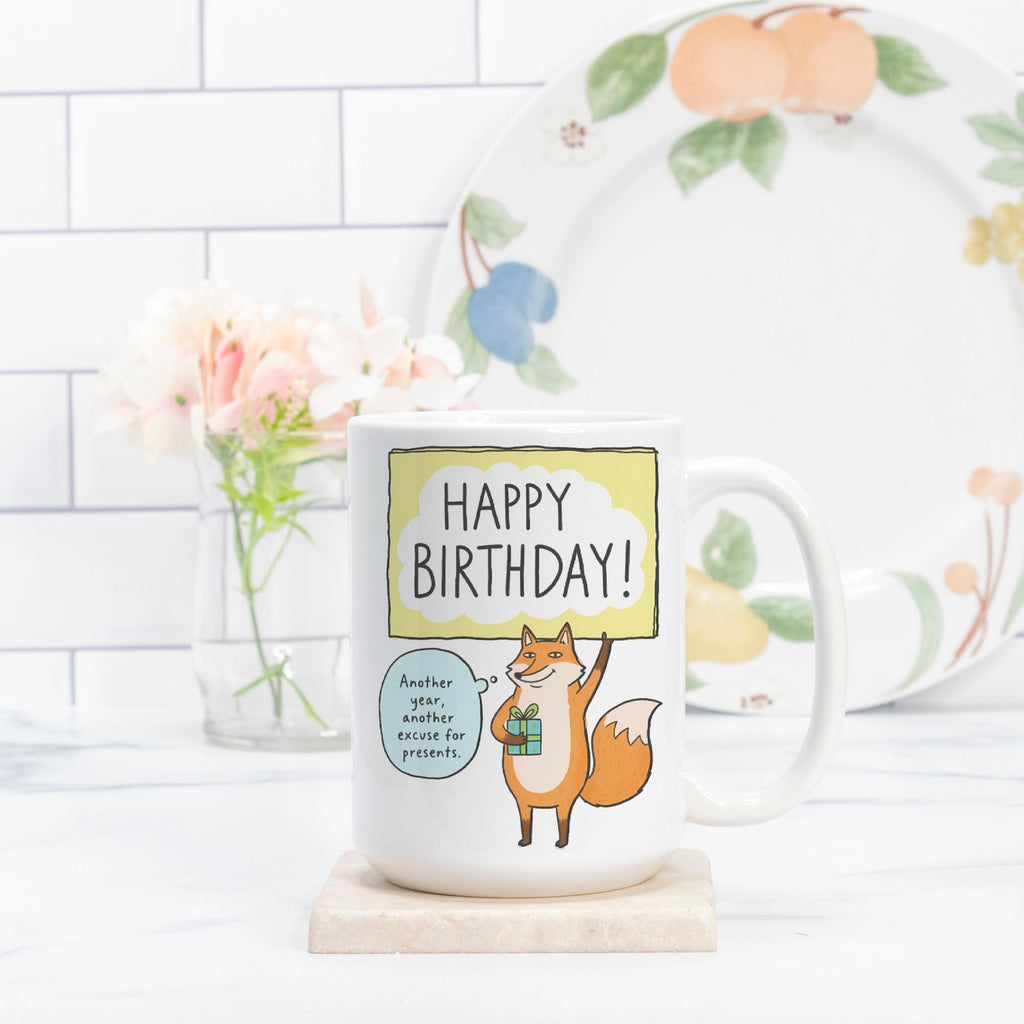 Mug with fox illustration and 'Happy Birthday!' text on a white surface with a floral plate in the background.
