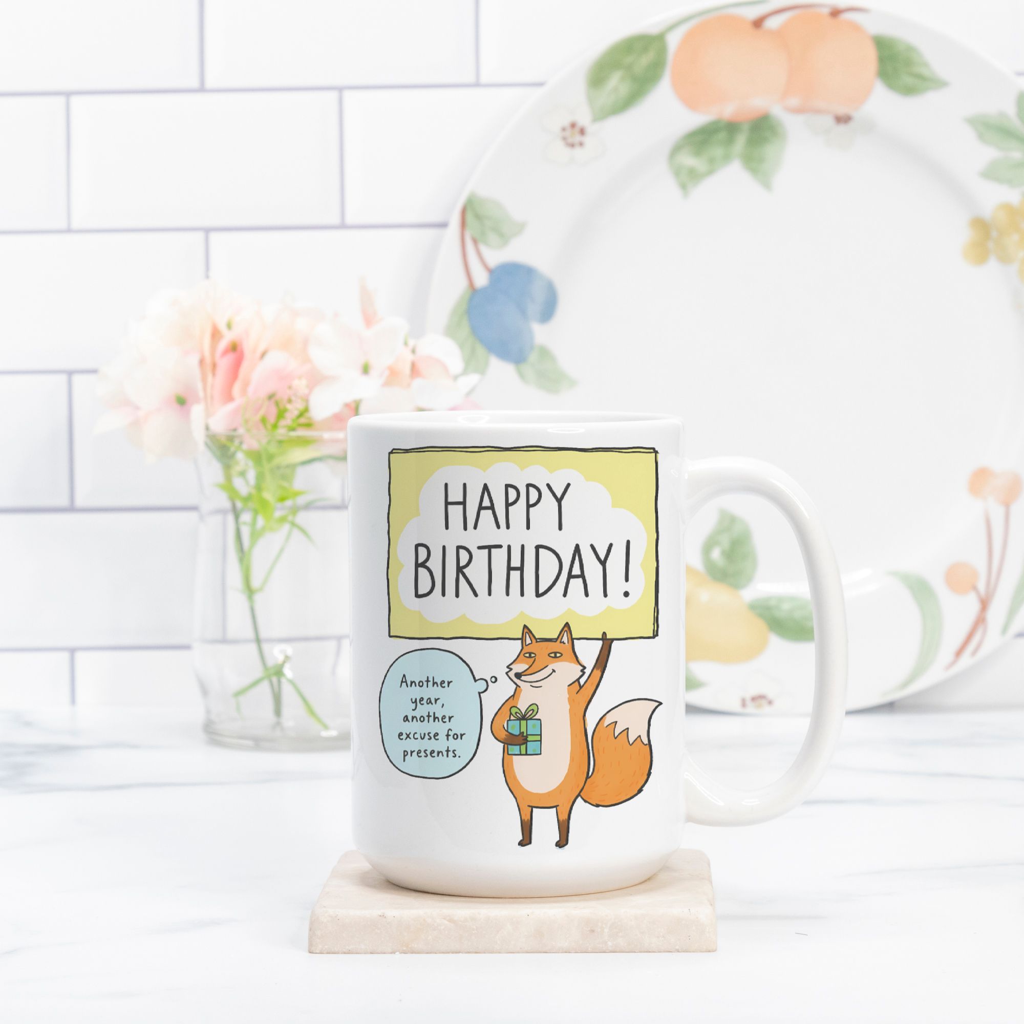 Mug with fox illustration and 'Happy Birthday!' text on a white surface with a floral plate in the background.