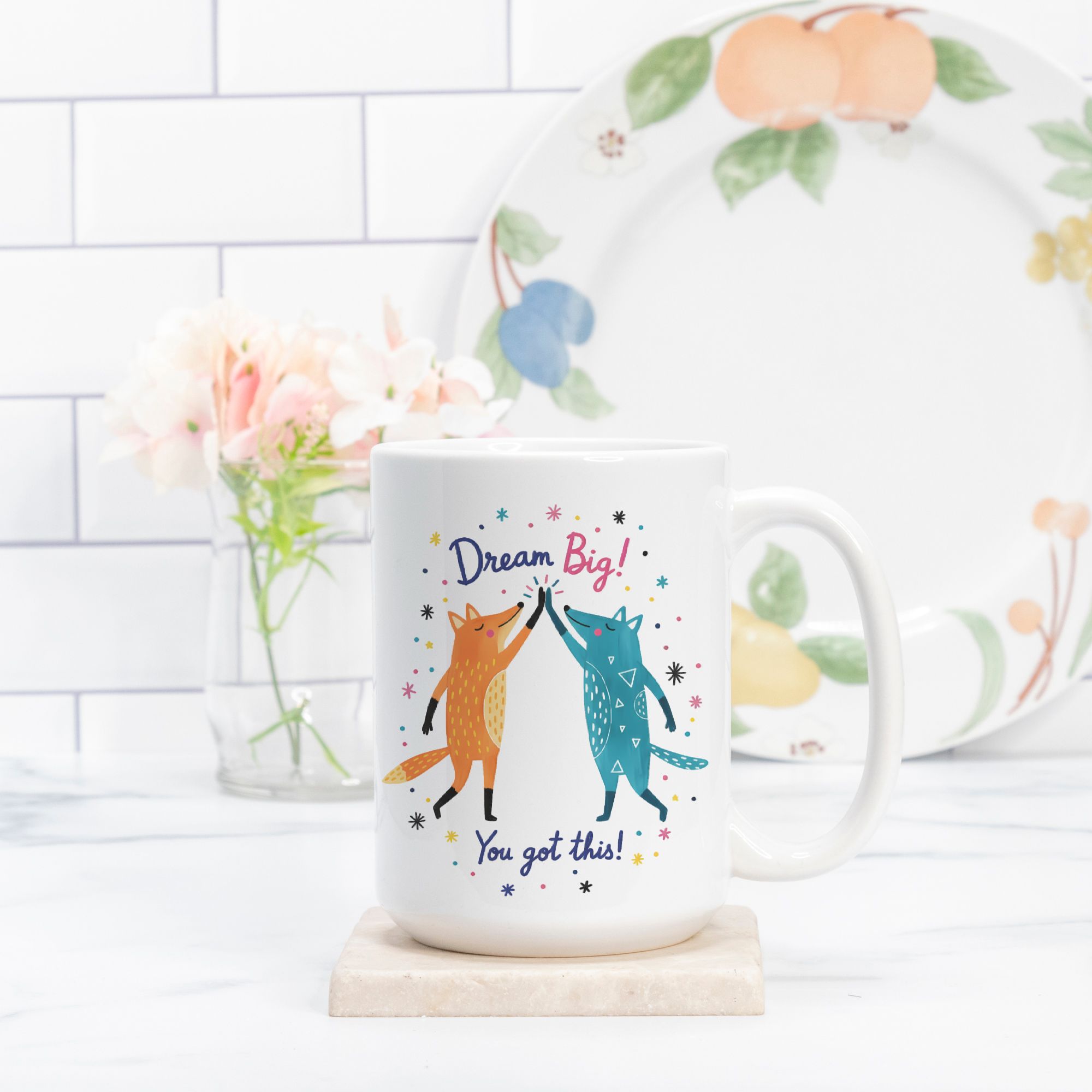 Mug with foxes and motivational quote on a white surface with a decorative plate in the background.
