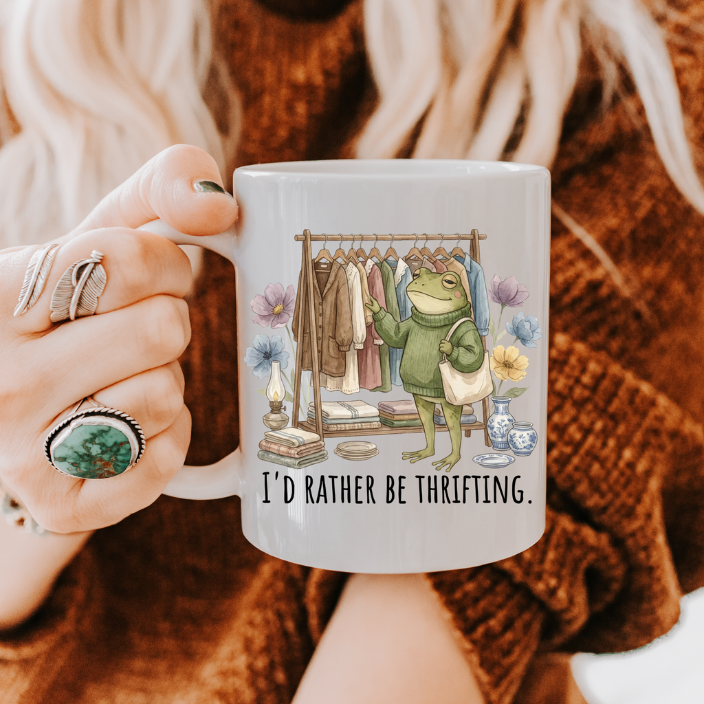 Person holding a mug with a frog graphic and text 'I'd rather be thrifting' against a textured brown background.