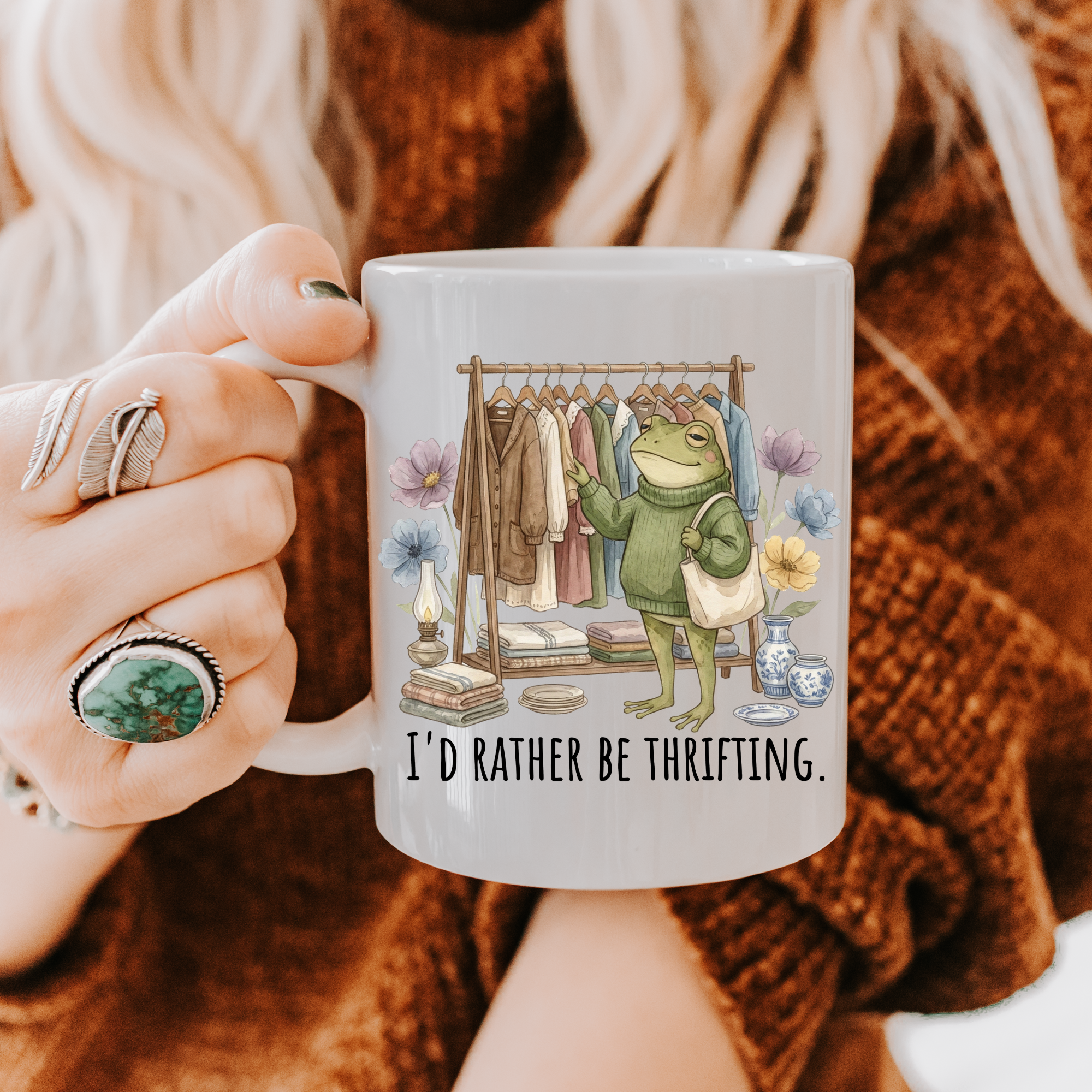 Person holding a mug with a frog graphic and text 'I'd rather be thrifting' against a textured brown background.