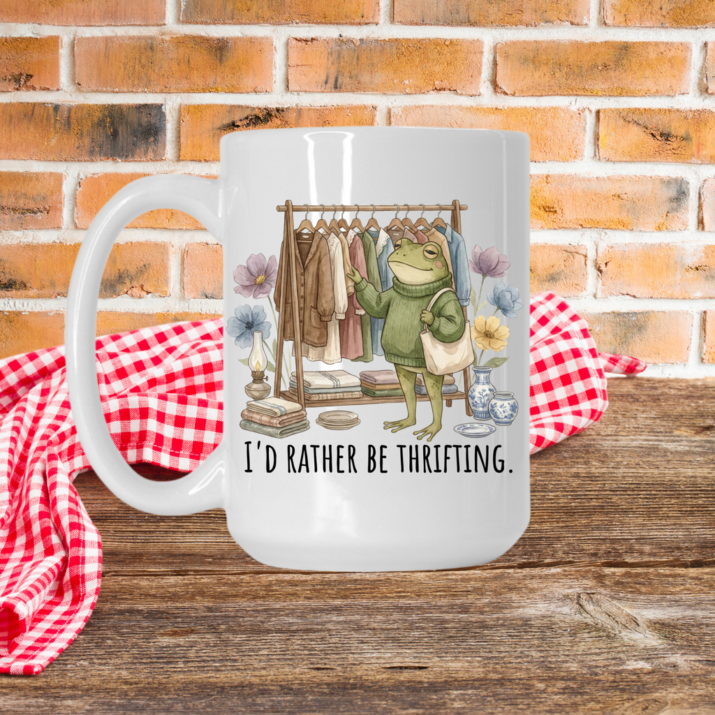 White 15oz. mug with a frog graphic and text 'I'd Rather Be Thrifting' on a wooden picnic table with a brick wall background.
