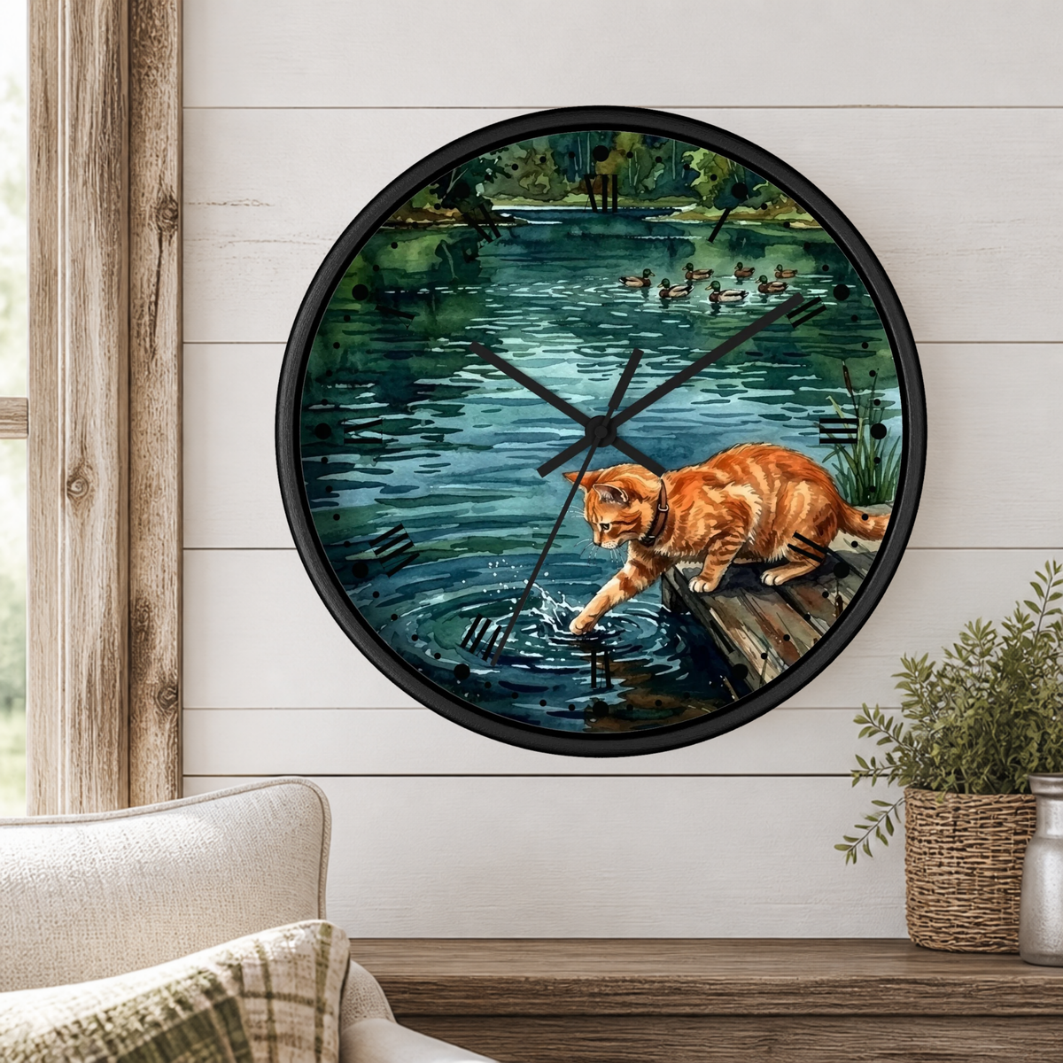 A round wall clock with a black frame hangs on a white shiplap wall above a wooden table. The clock face features a painted scene of an orange tabby cat standing on a log by a lake, with ducks and trees in the background.