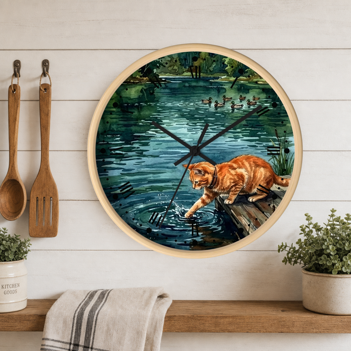 Round wall clock with a light wood frame featuring a painted scene of an orange tabby cat at the edge of a pond, displayed on a white wood plank wall above a shelf with plants and kitchen utensils.