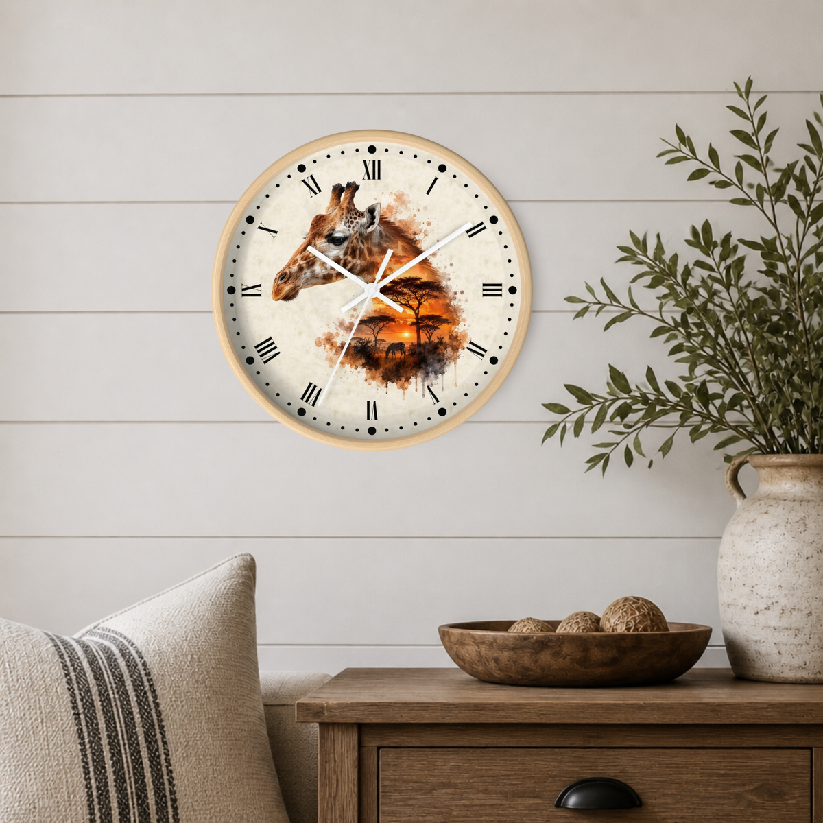A round wall clock with a giraffe and African savanna watercolor design on the face, mounted on a white shiplap wall above a wooden side table with a plant and decorative bowl.