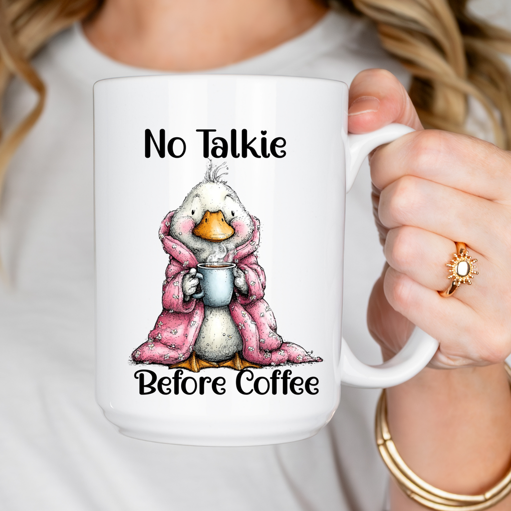 Mug with a cartoon goose in a pink robe holding a coffee cup, text 'No Talkie Before Coffee', held by a person.