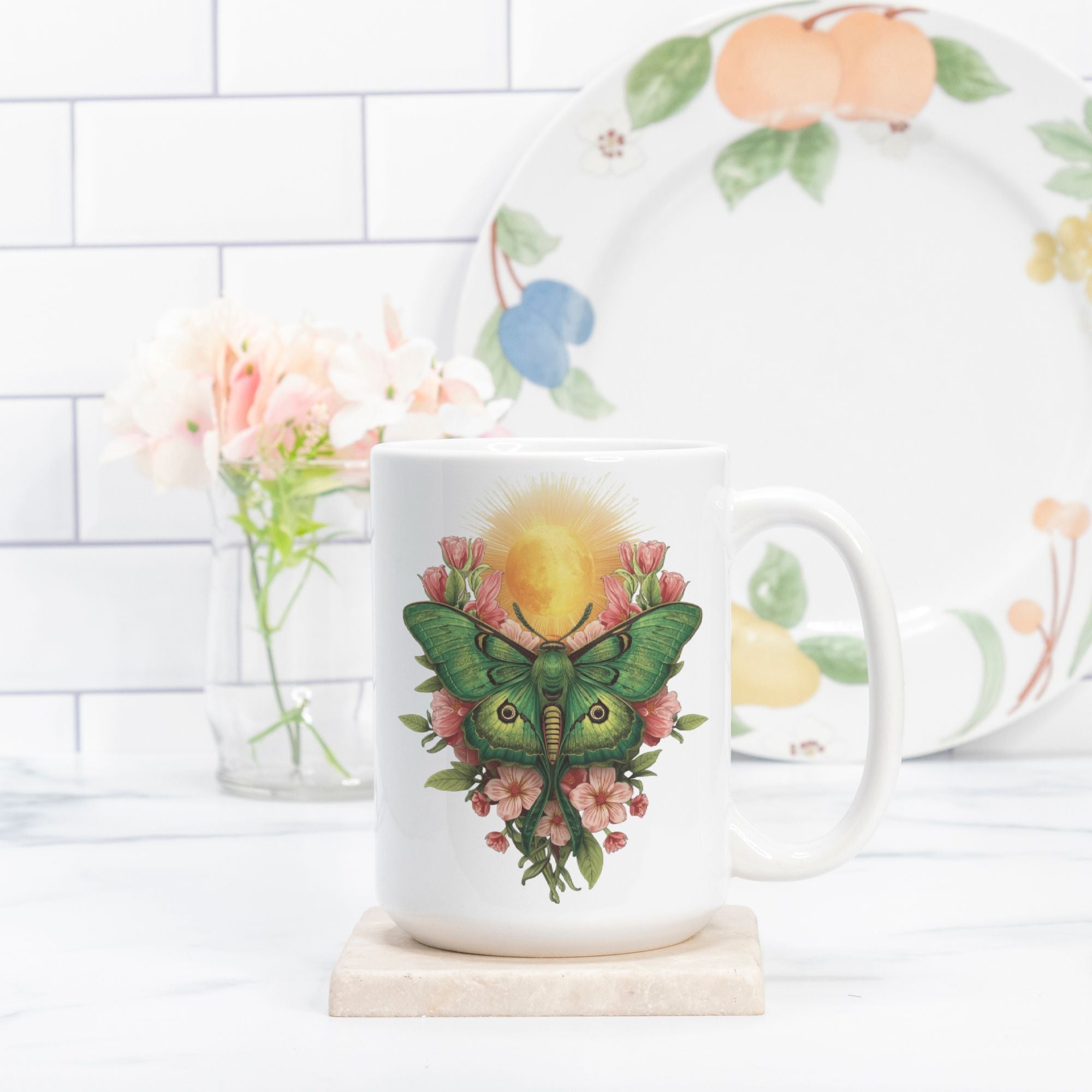 White mug with a green lunar moth design on a white surface with a floral plate in the background.