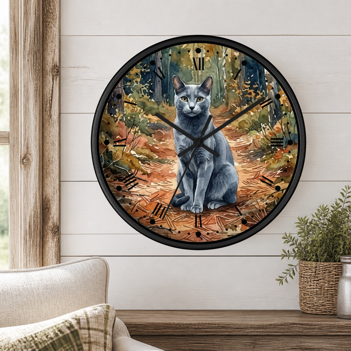 A round wall clock with a black frame and a printed watercolor illustration of a gray Russian blue cat sitting in an autumn forest, mounted on a white shiplap wall above a wooden mantel with a plant and cushion.