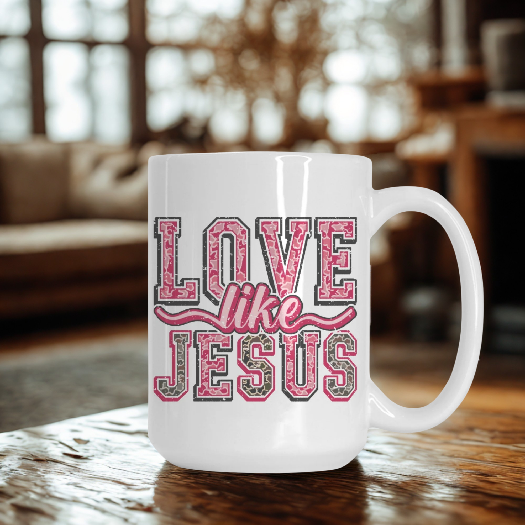 White mug with pink and grey camo lettering 'Love Like Jesus' on a wooden table in a living room.