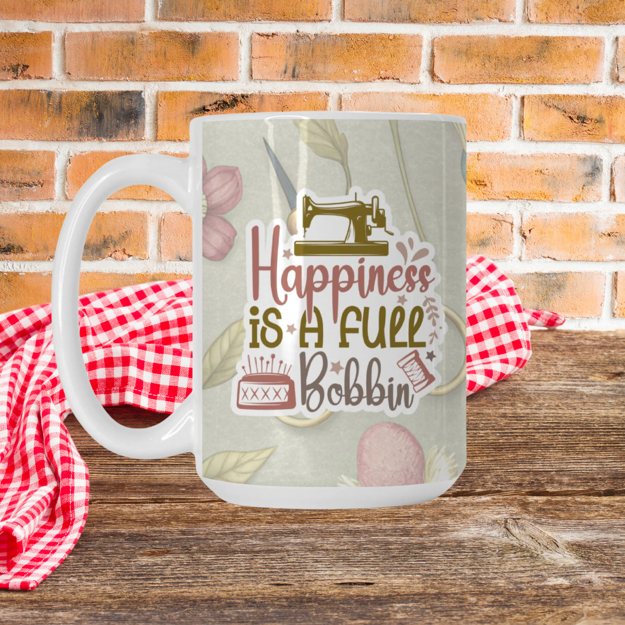 Mug with sewing-themed design and text 'Happiness is a Full Bobbin' on a wooden surface with a red and white checkered cloth and brick wall background.