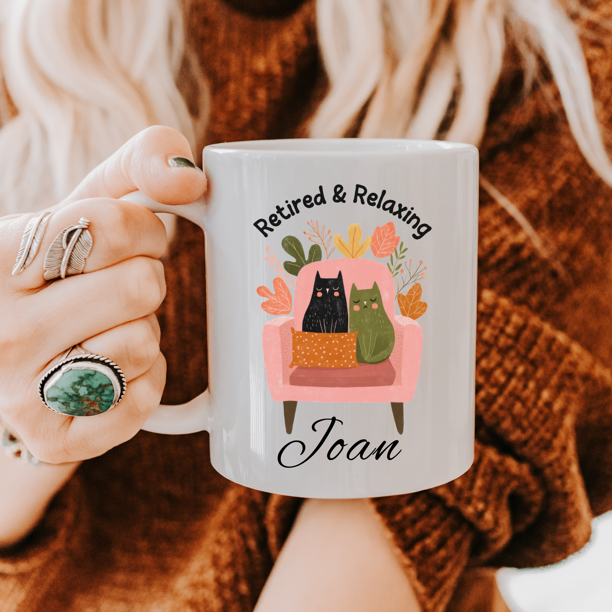 Person holding an 11oz. mug with a design of two cats on a couch and text 'Retired & Relaxing Joan'.
