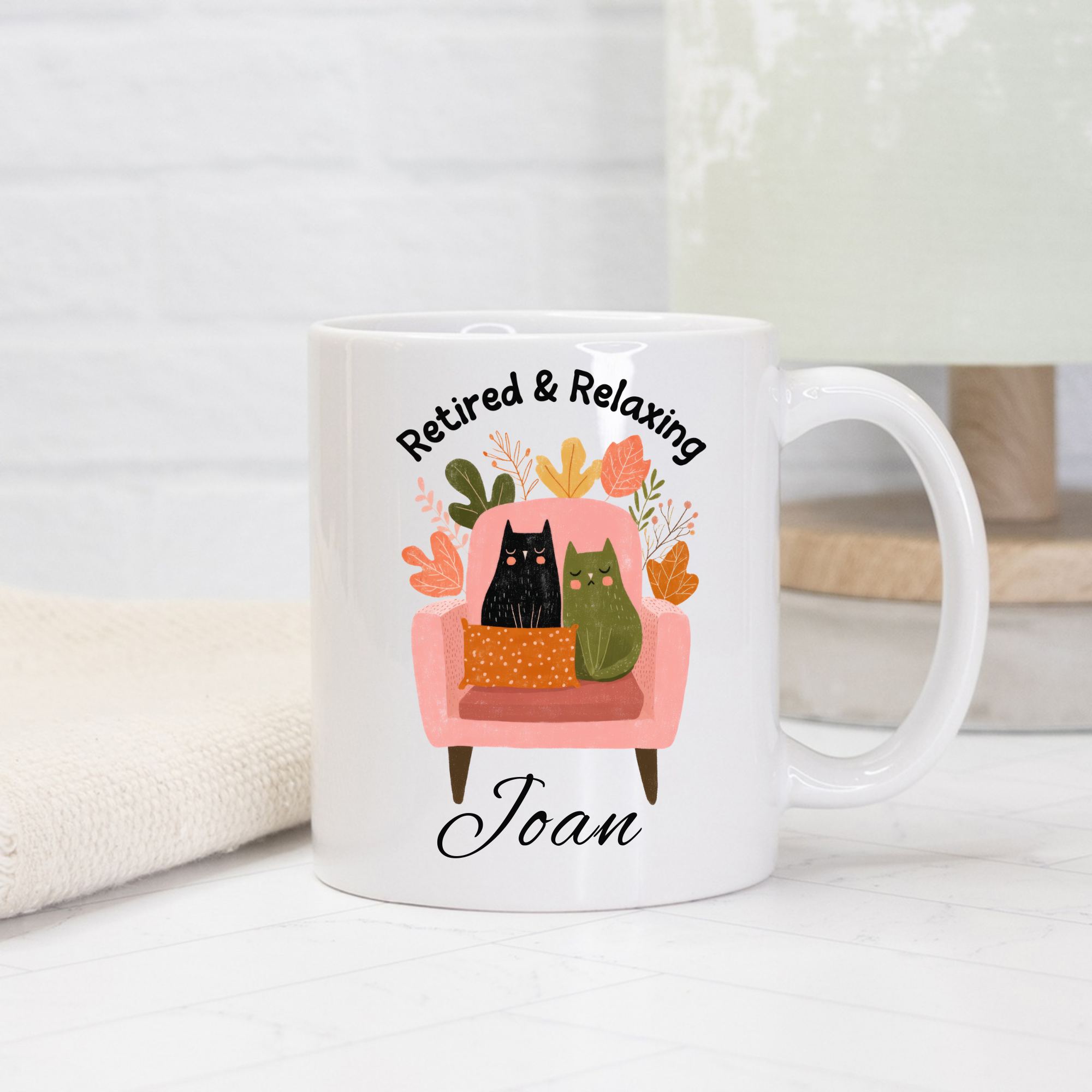 White 11oz. mug with a design of two cats on a pink chair, text 'Retired & Relaxing Joan', and decorative elements.