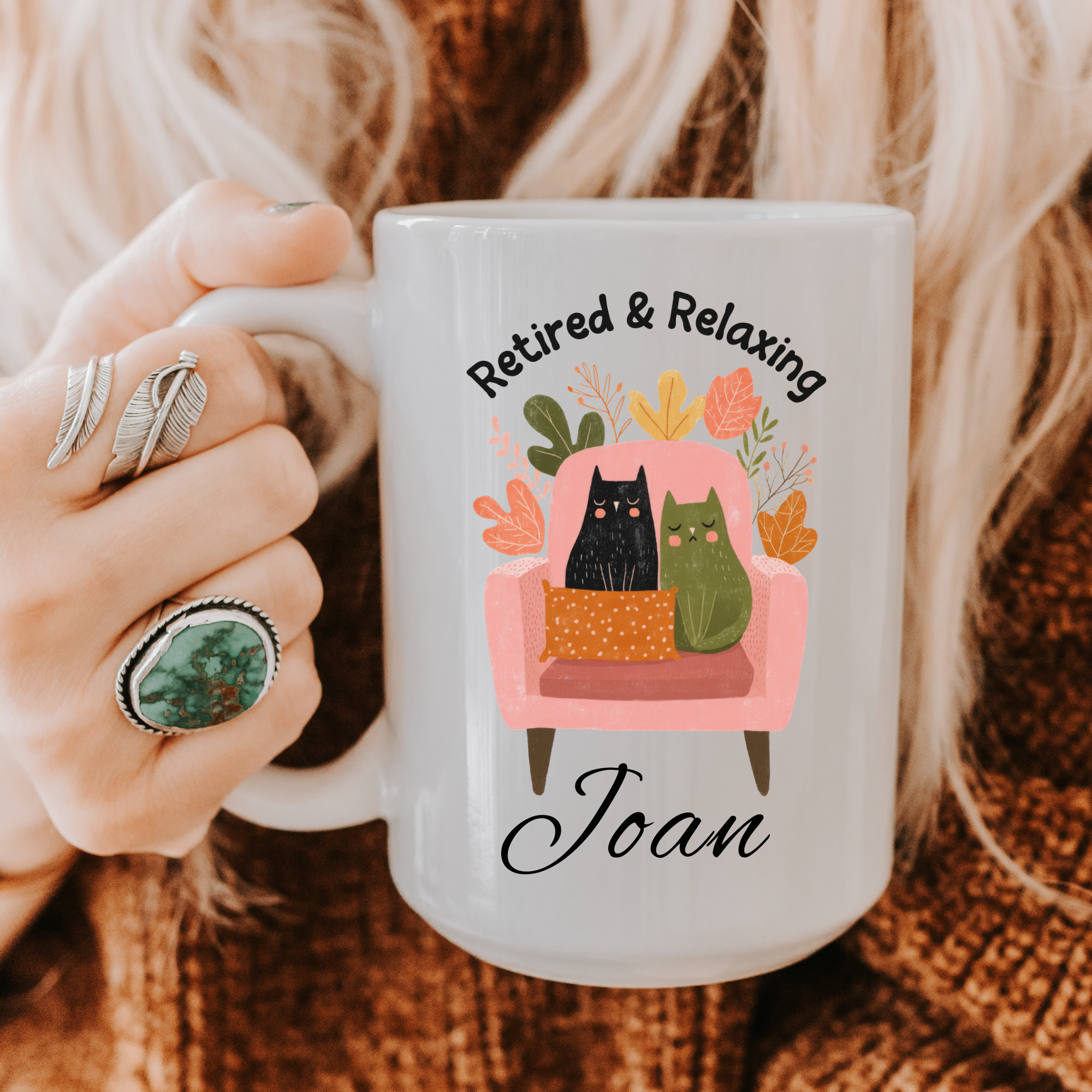 Person holding a mug with 'Retired & Relaxing Joan' design featuring cats on an armchair.