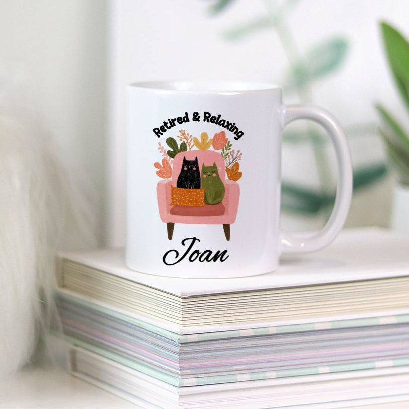 White mug with a design of cats on a chair and text 'Retired & Relaxing Joan' on a stack of books.