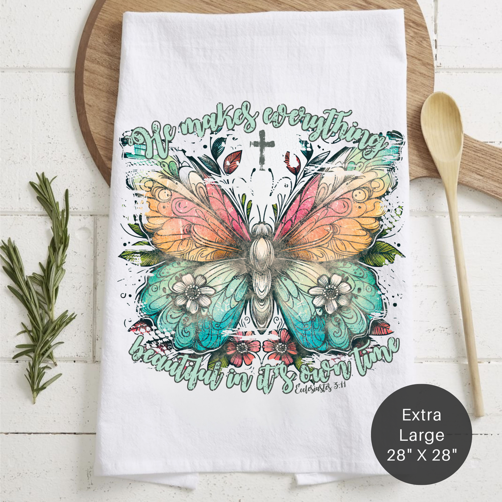 White tea towel with colorful butterfly design and text 'He Makes Everything Beautiful In His Own Time' on a wooden surface.