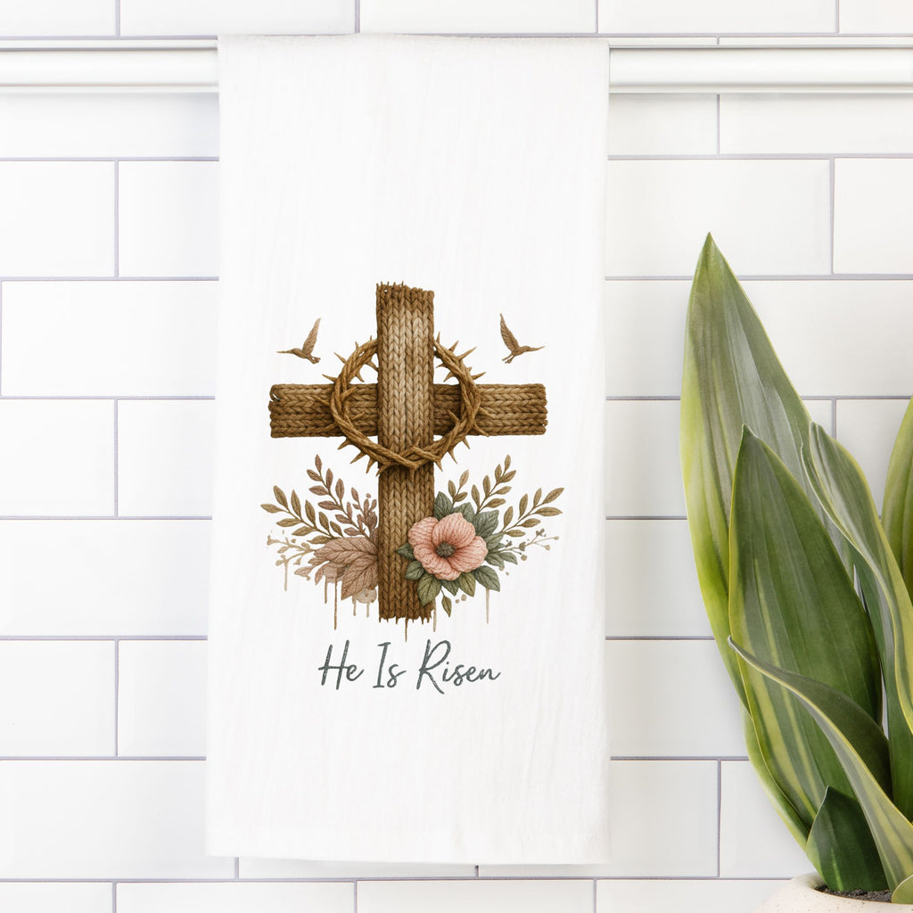 Decorative tea towel with cross,  floral elements, and 'He Is Risen' text on a white tiled wall.