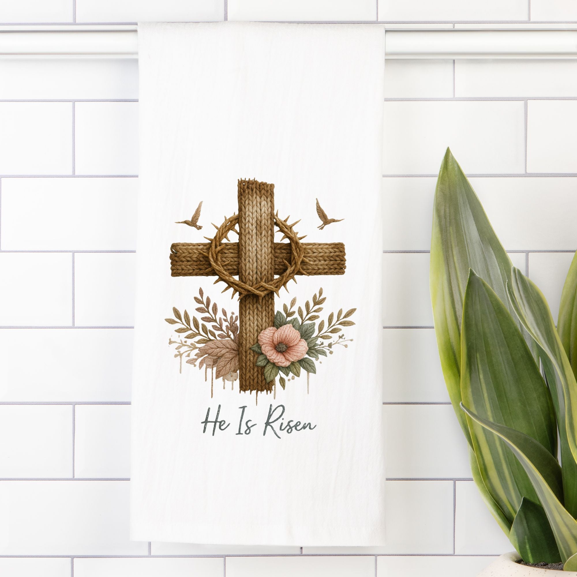 Decorative tea towel with cross,  floral elements, and 'He Is Risen' text on a white tiled wall.