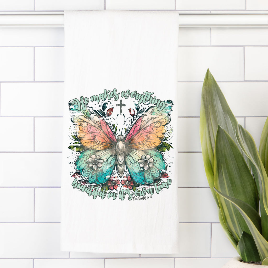 Tea towel with colorful butterfly design with text on a white tiled wall, next to a plant.