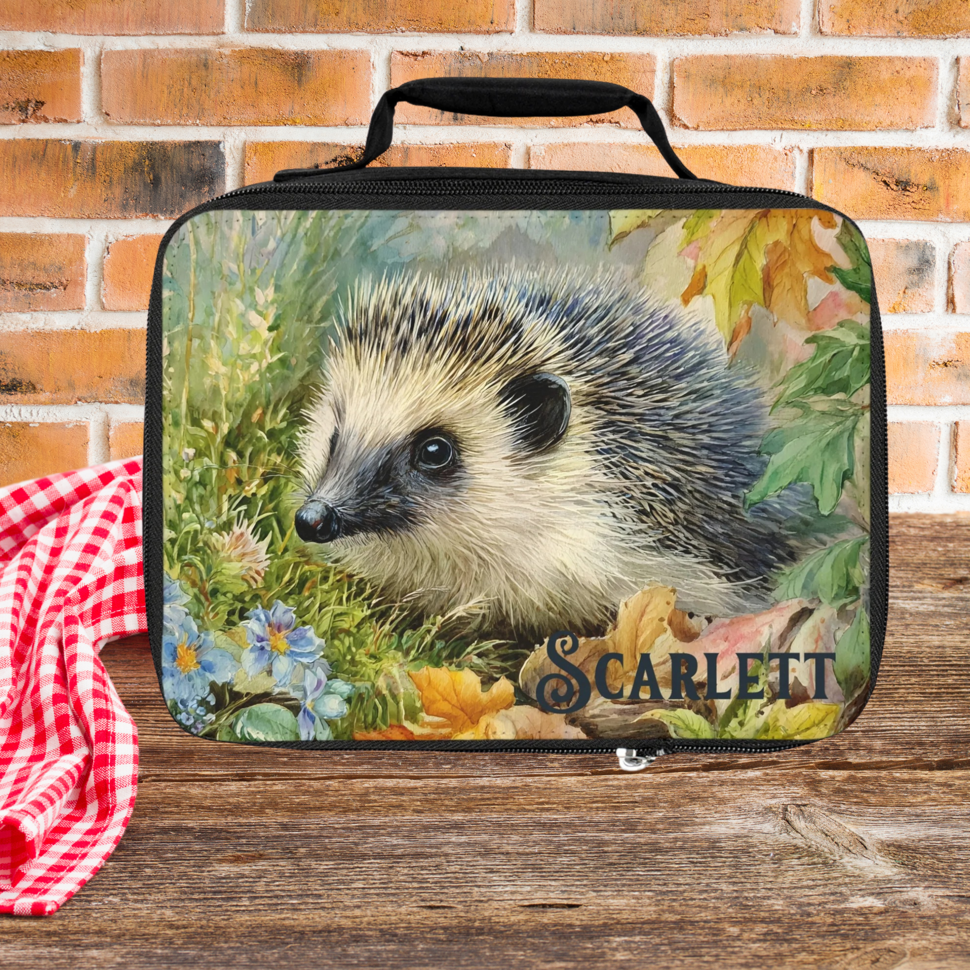 Lunch bag with hedgehog design and 'Scarlett' custom text on a wooden picnic table with brick background