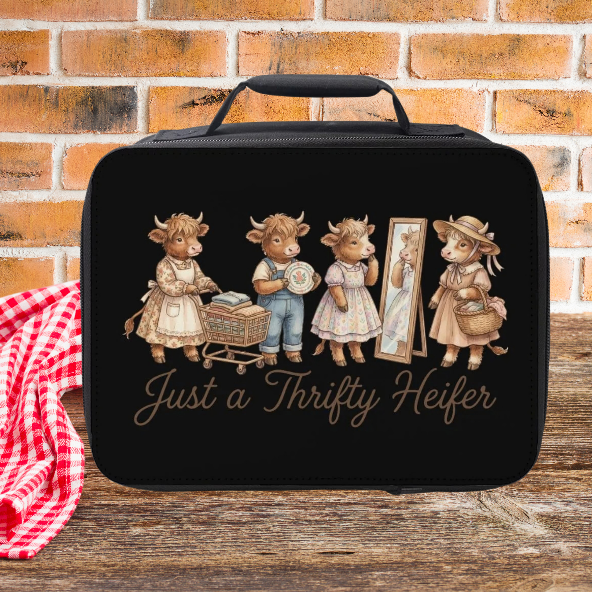 Black lunch bag with cow illustrations and text 'Just a Thrifty Heifer' on a wooden picnic table with a brick wall background.