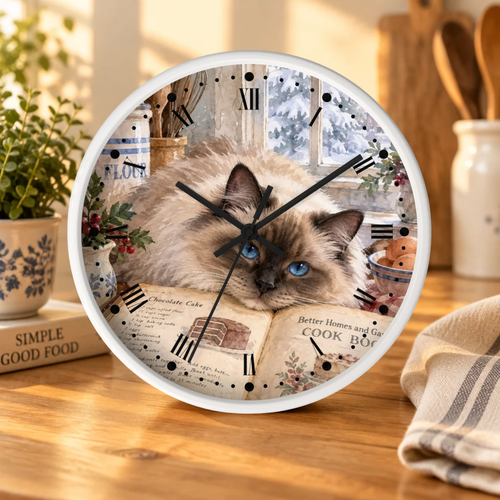 A round wall clock with a white frame sits on a wooden table. The clock face features a photo of a fluffy cream-colored cat with dark brown patches and bright blue eyes, surrounded by vintage kitchen illustrations, Roman numerals, and black clock hands.