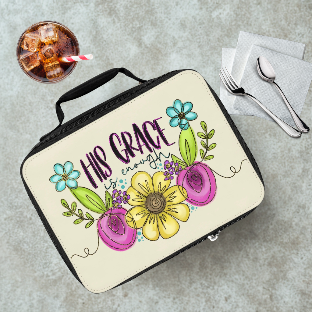 Lunch box with floral design and 'His Grace is Enough' text on a cafeteria table with a drink and utensils.