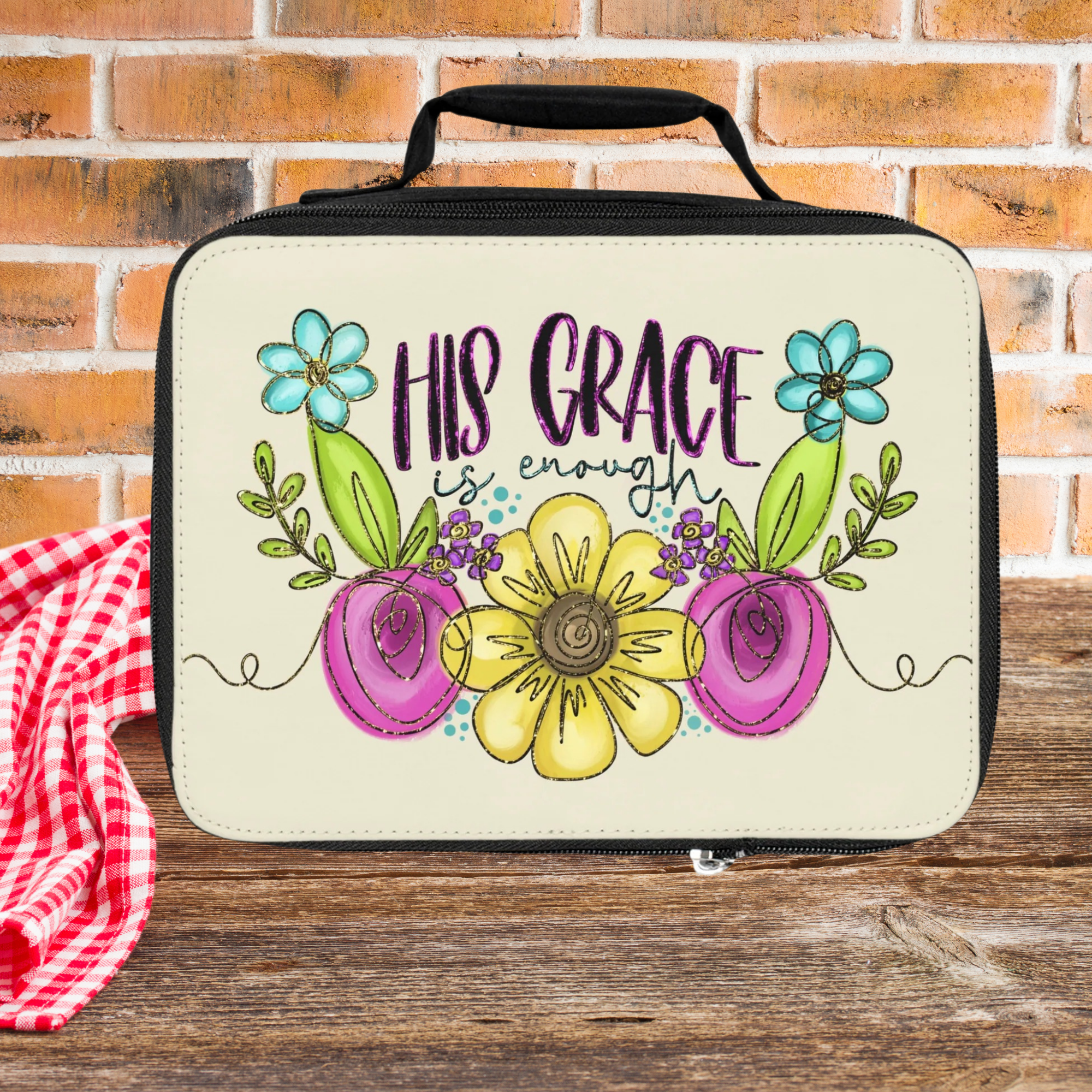Lunch bag with floral design and 'His Grace is enough' text on a wooden picnic table against a brick wall background