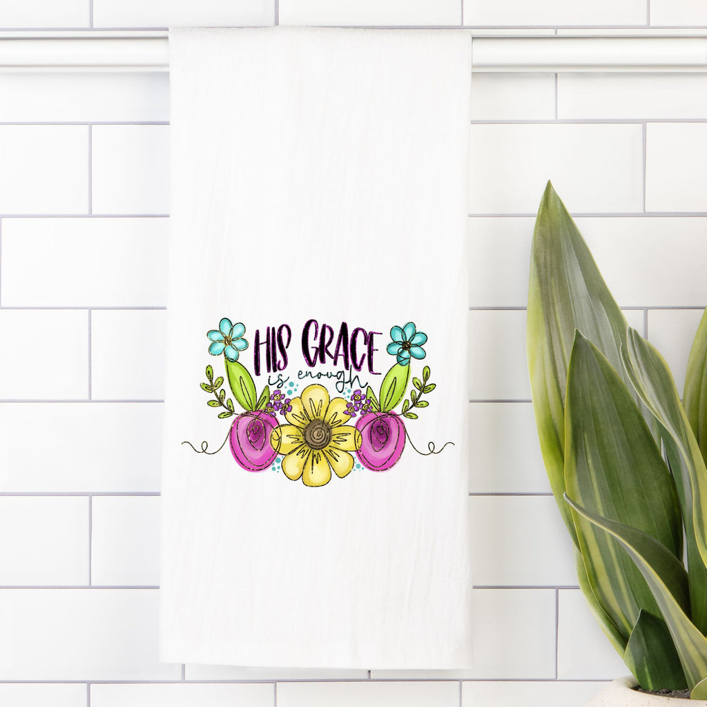 Decorative tea towel with floral design and 'His Grace is Enough' text on a white tiled wall.