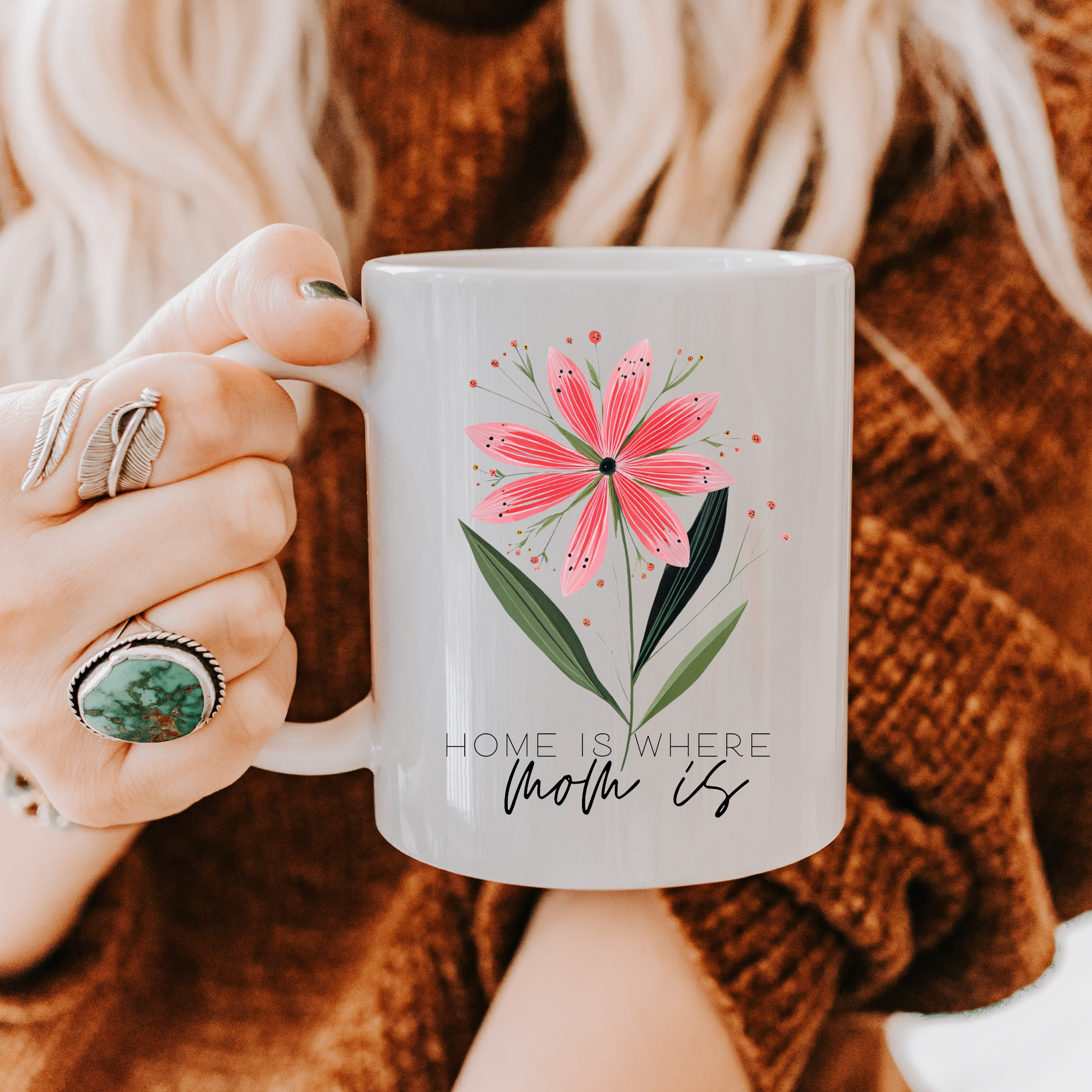 Person holding an 11oz white ceramic mug with a floral design and text 'Home Is Where Mom Is', wearing a brown sweater.